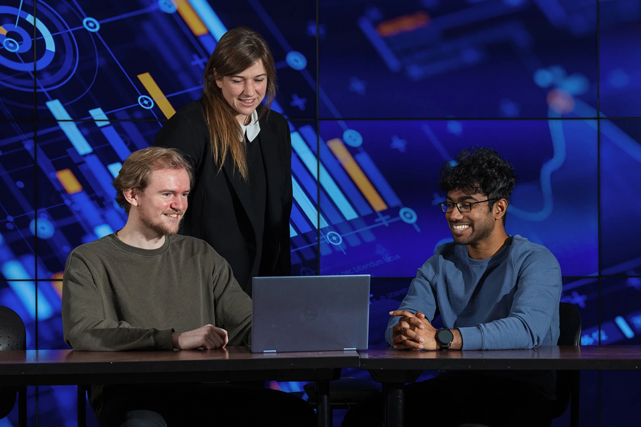 Three students viewing a laptop screen with a wall of LCD panels showing an abstract graphic behind them